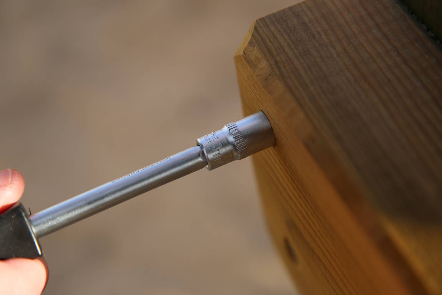 Close-up van een moersleutel die in een houten balk wordt geschroefd. Een menselijke hand houdt de greep van het gereedschap vast.