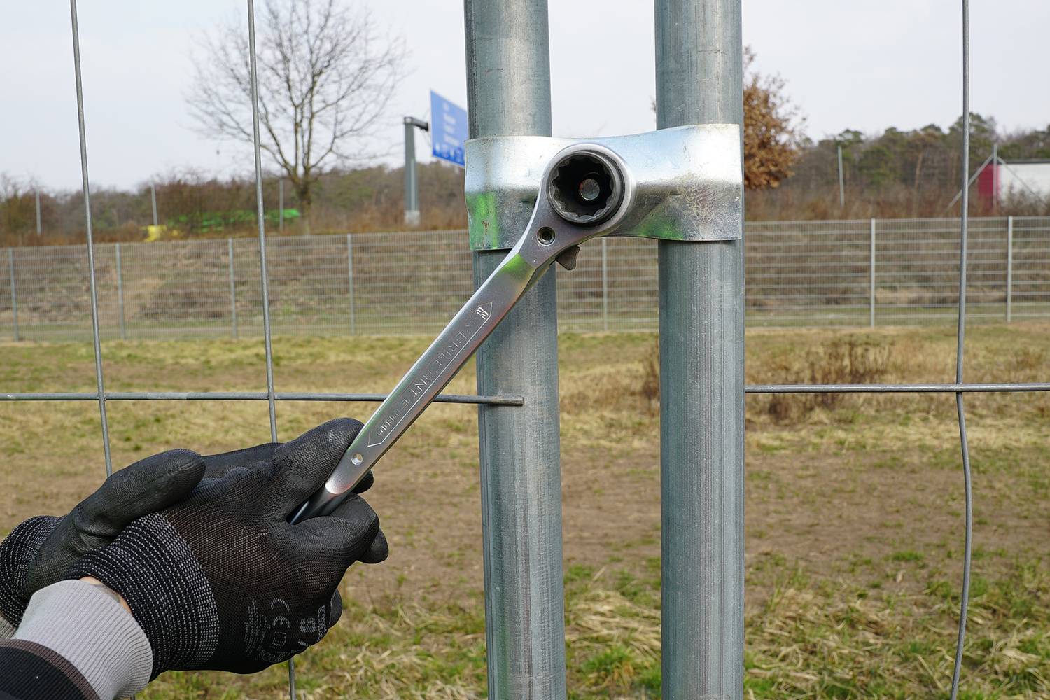 Een persoon draait met een moersleutel een schroef aan die twee metalen buizen bij elkaar houdt. Op de achtergrond is een hek zichtbaar.