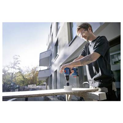 Een persoon gebruikt een elektrische boormachine om een gat te maken in een betonblok, draagt een veiligheidsbril, buiten een modern gebouw.