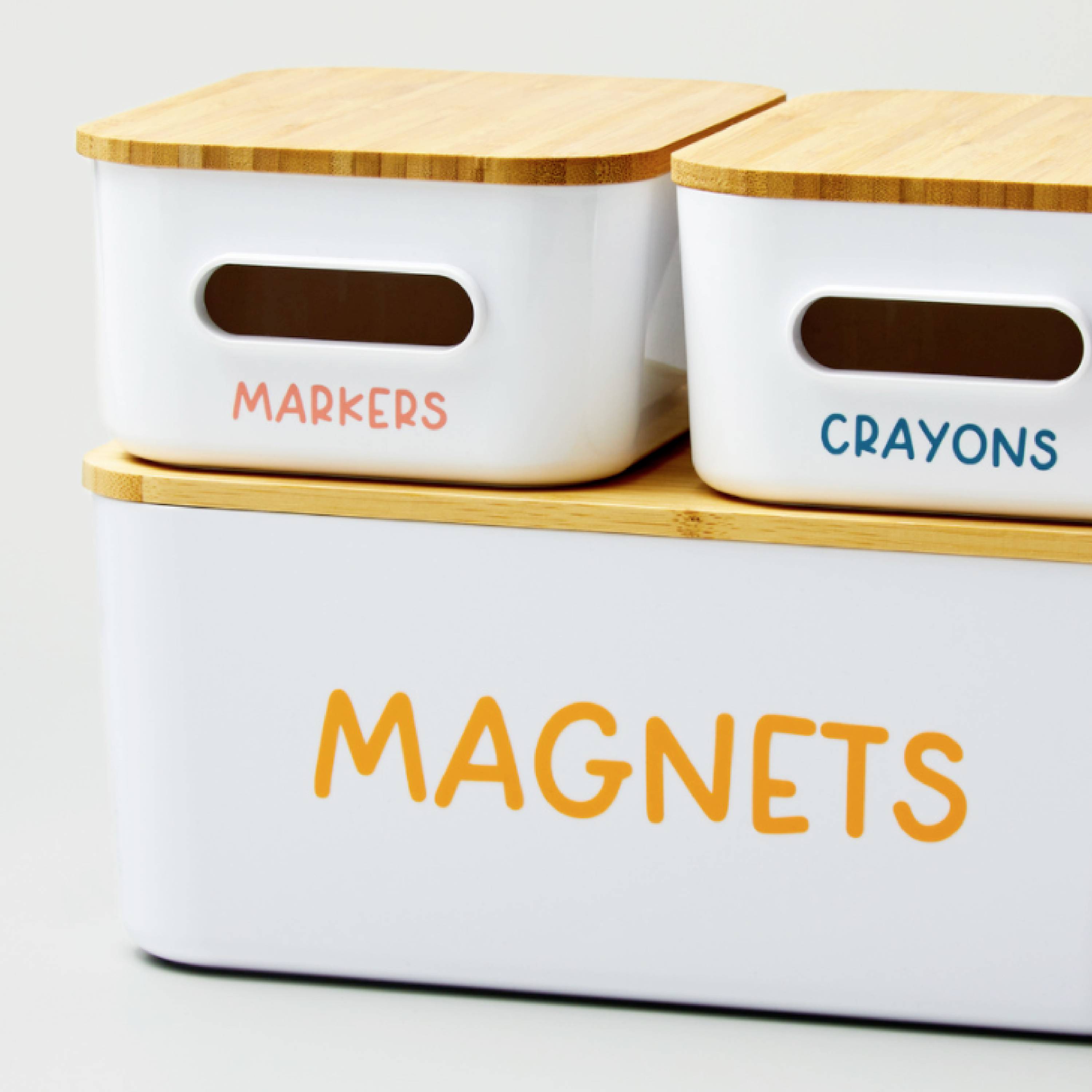 Drie witte containers met houten deksels, gelabeld met 'Markers', 'Crayons' en 'Magnets', ter organisatie van kantoorartikelen.