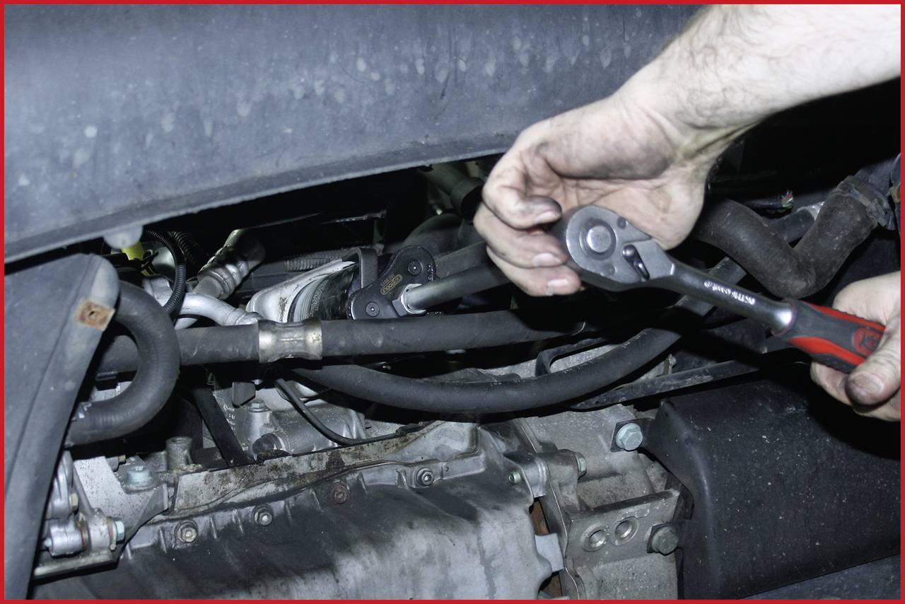 Een monteur met handschoenen repareert de motor van een voertuig met een moersleutel. Hij werkt aan leidingen en kabels.