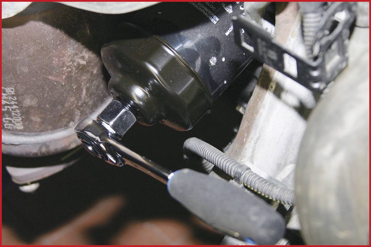 Close-up van een oliefilter in de motorruimte van een auto, waarbij deze wordt losgedraaid met een moersleutel. Mechanische details zijn duidelijk zichtbaar.
