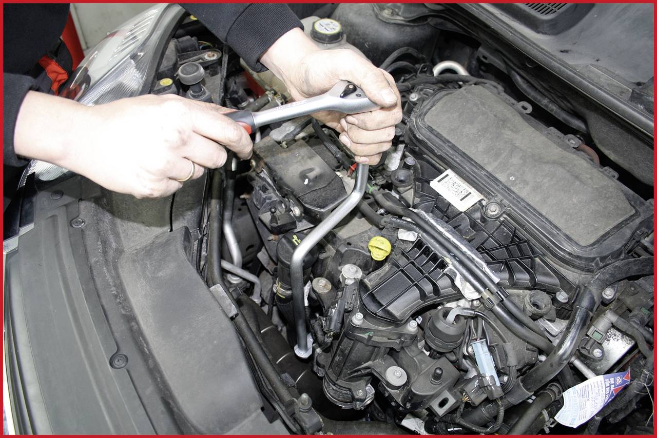 Een persoon repareert de motor van een auto met een moersleutel. Verschillende leidingen en componenten zijn zichtbaar.