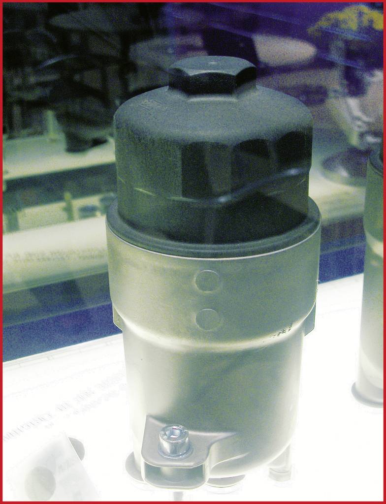 Metalen cilinder met zwarte afdekking, mogelijk een filter of mechanisch onderdeel, geplaatst in een tentoonstellingsvitrine.