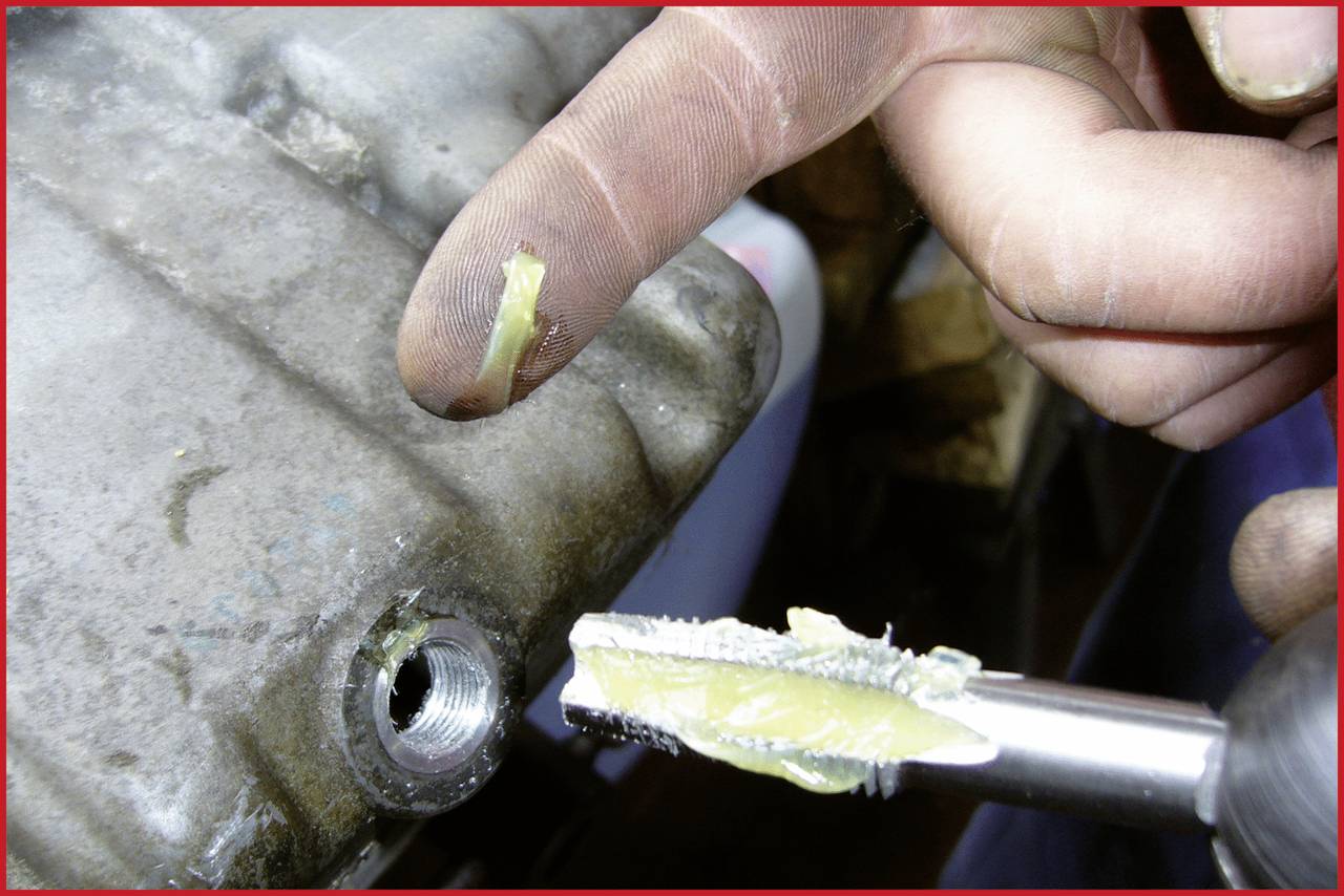 Een persoon brengt vet aan op de punt van een gereedschap met zijn vinger. Een metalen onderdeel met een schroefdraadgat is zichtbaar.