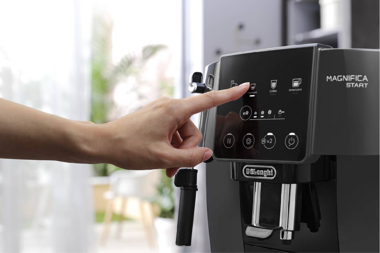 Een hand bedient het touchscreen van een zwarte koffiemachine om het koffiezetten te starten. Op de achtergrond zijn onscherpe planten zichtbaar.