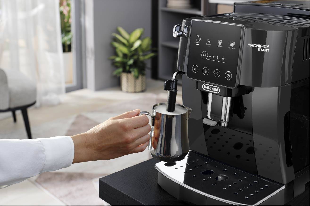 Een persoon schuimt melk met een espressomachine. De keuken is modern ingericht, met planten op de achtergrond.
