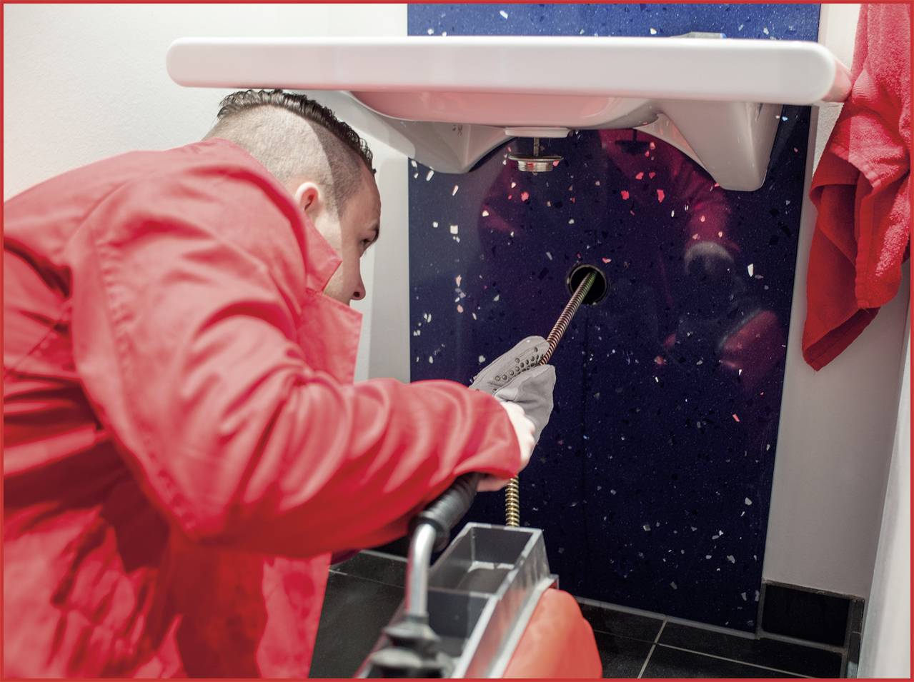 Een loodgieter in rode werkkleding repareert de afvoerpijp onder een wit wastafel.