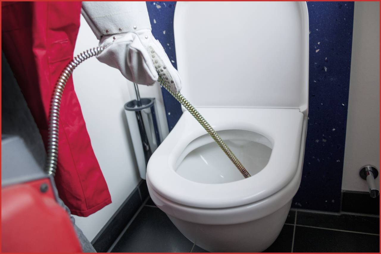 Een persoon in werkkleding gebruikt een buizenreinigingsspiraal om een toilet te reinigen. De focus ligt op het reinigingsproces.
