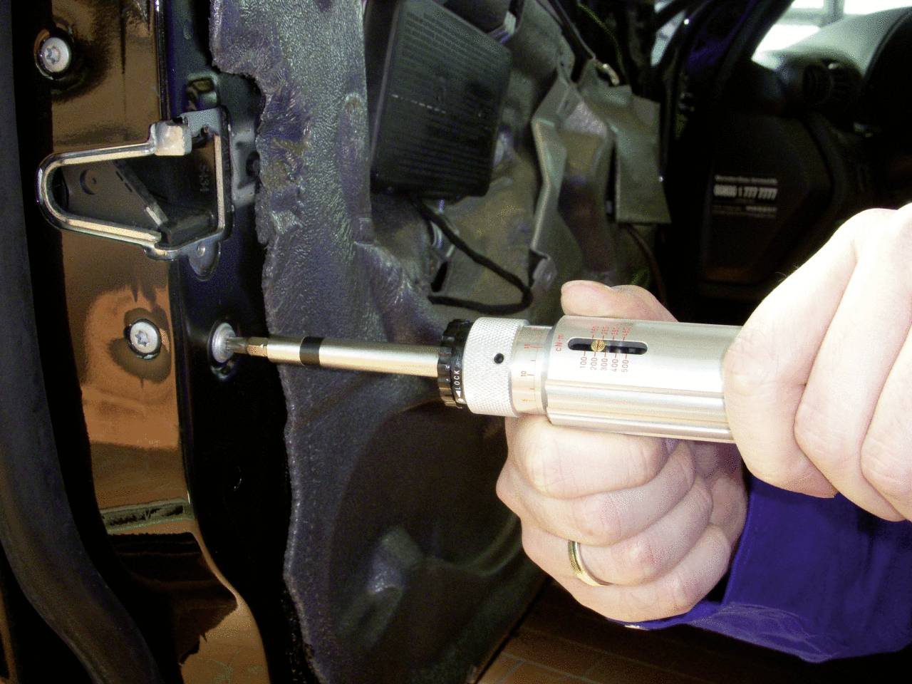 Een persoon bevestigt met een schroevendraaier een schroef aan een autoportier. De omgeving doet vermoeden dat dit in een werkplaats plaatsvindt.