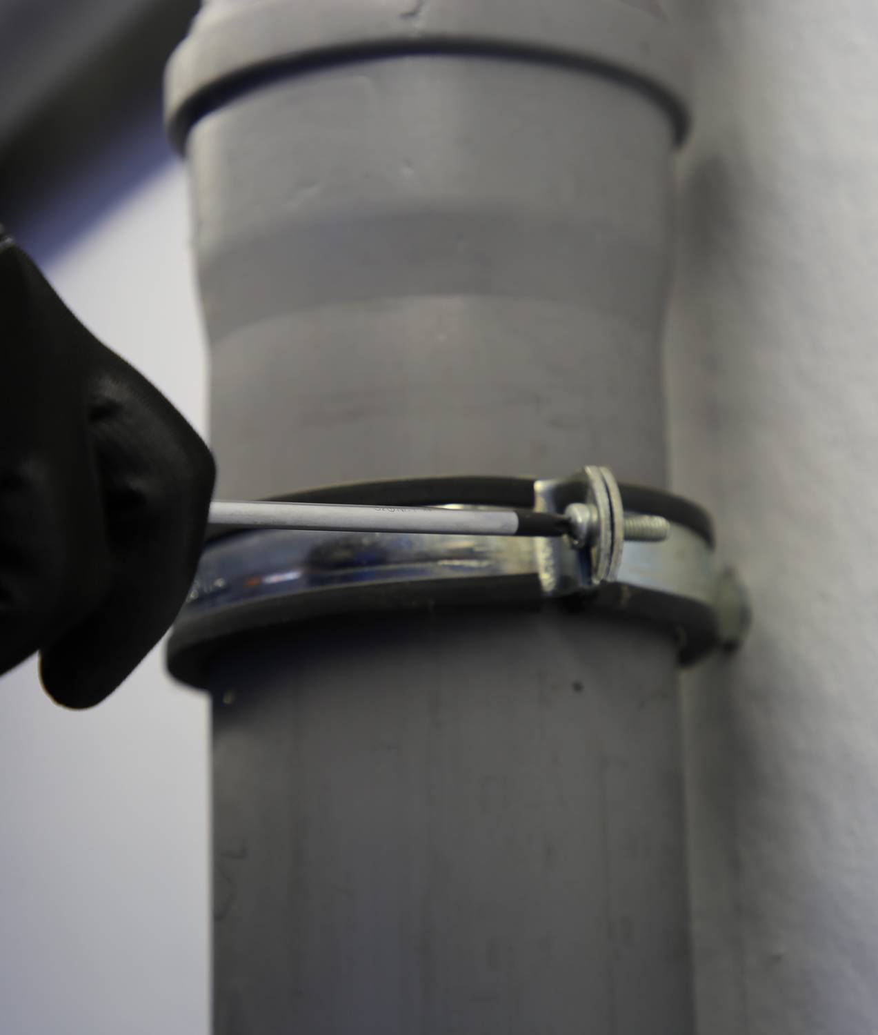 Een vakman draait een schroef vast aan een buisklem. De buis is groot en grijs, de hand draagt een zwarte handschoen.