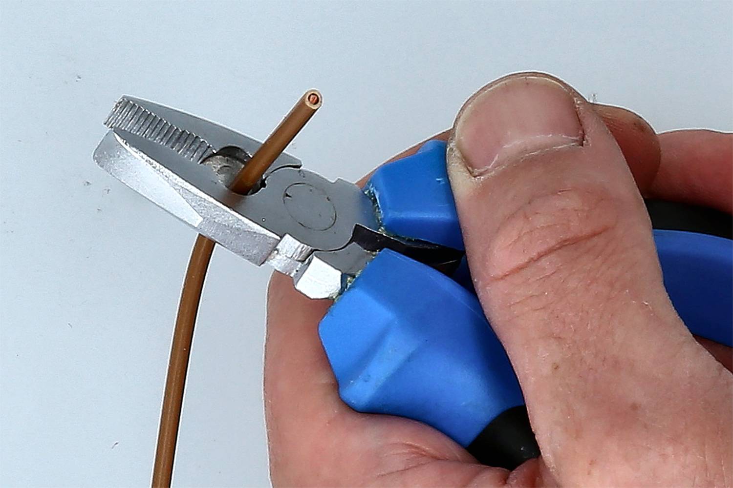 Een hand houdt een tang vast die een bruine kabel doorsnijdt. De tang heeft blauwe handgrepen.
