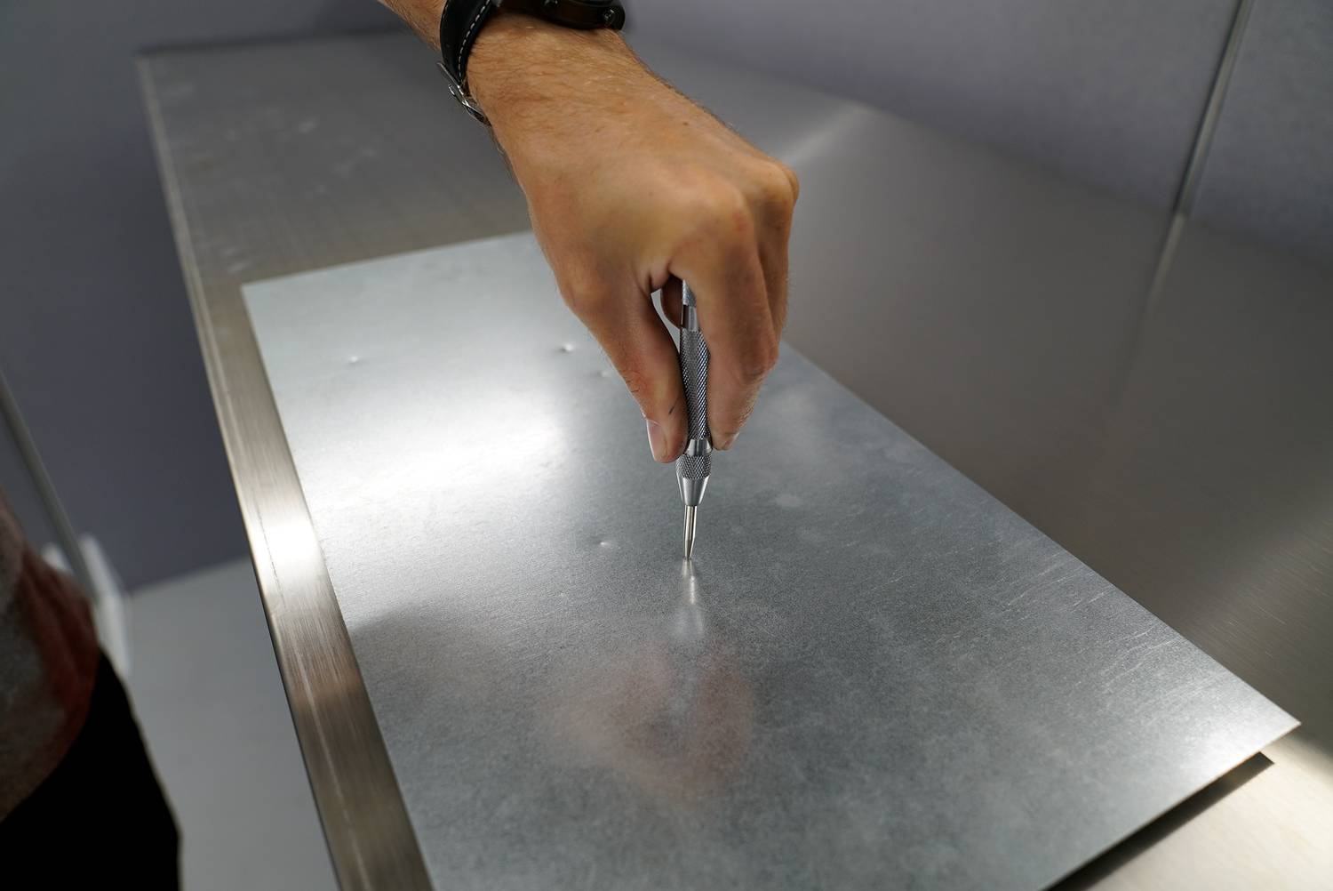 Een hand houdt een gereedschap vast boven een metalen plaat en zet een punt. De scène toont een werkstap in de metaalbewerking.