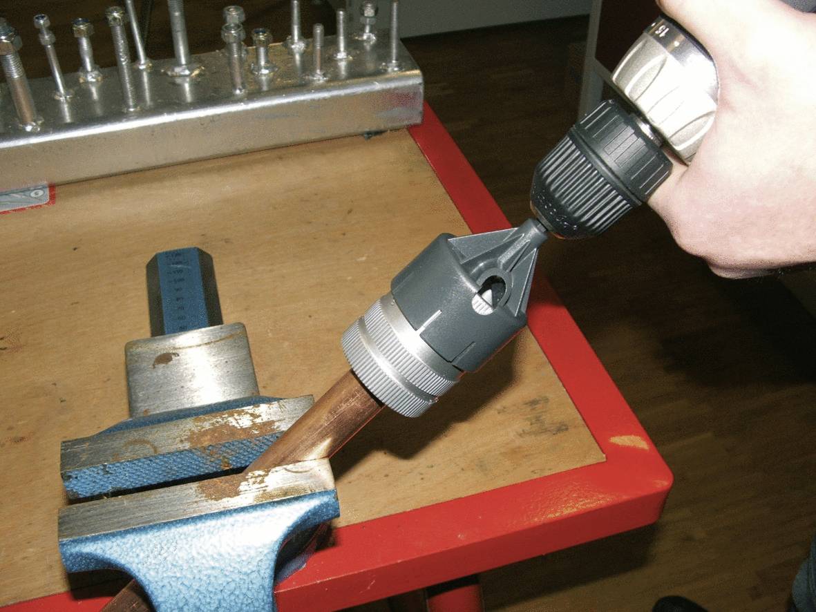 Een persoon gebruikt een boormachine om een koperen buis te ontbramen in een bankschroef. Op de achtergrond zijn gereedschappen zichtbaar.