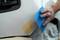 Een hand brengt gele plamuur aan op een witte autocarrosserie. De plamuur wordt verdeeld met een blauw plastic gereedschap.
