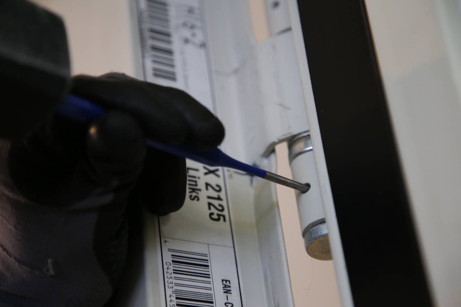 Een persoon met een zwarte handschoen draait met een gereedschap een schroef vast aan een scharnier, naast een etiket met tekst.