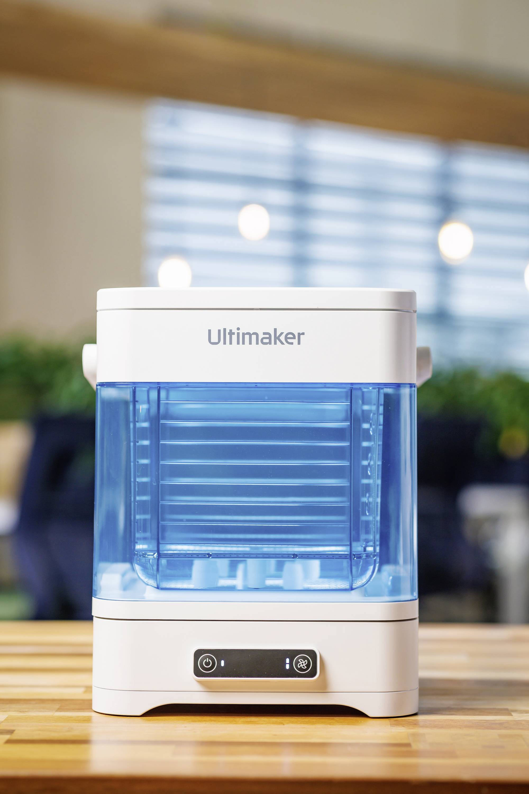 3D-printer met een transparante blauwe behuizing en een witte deksel, staand op een houten tafel, met 'Ultimaker'-opschrift.