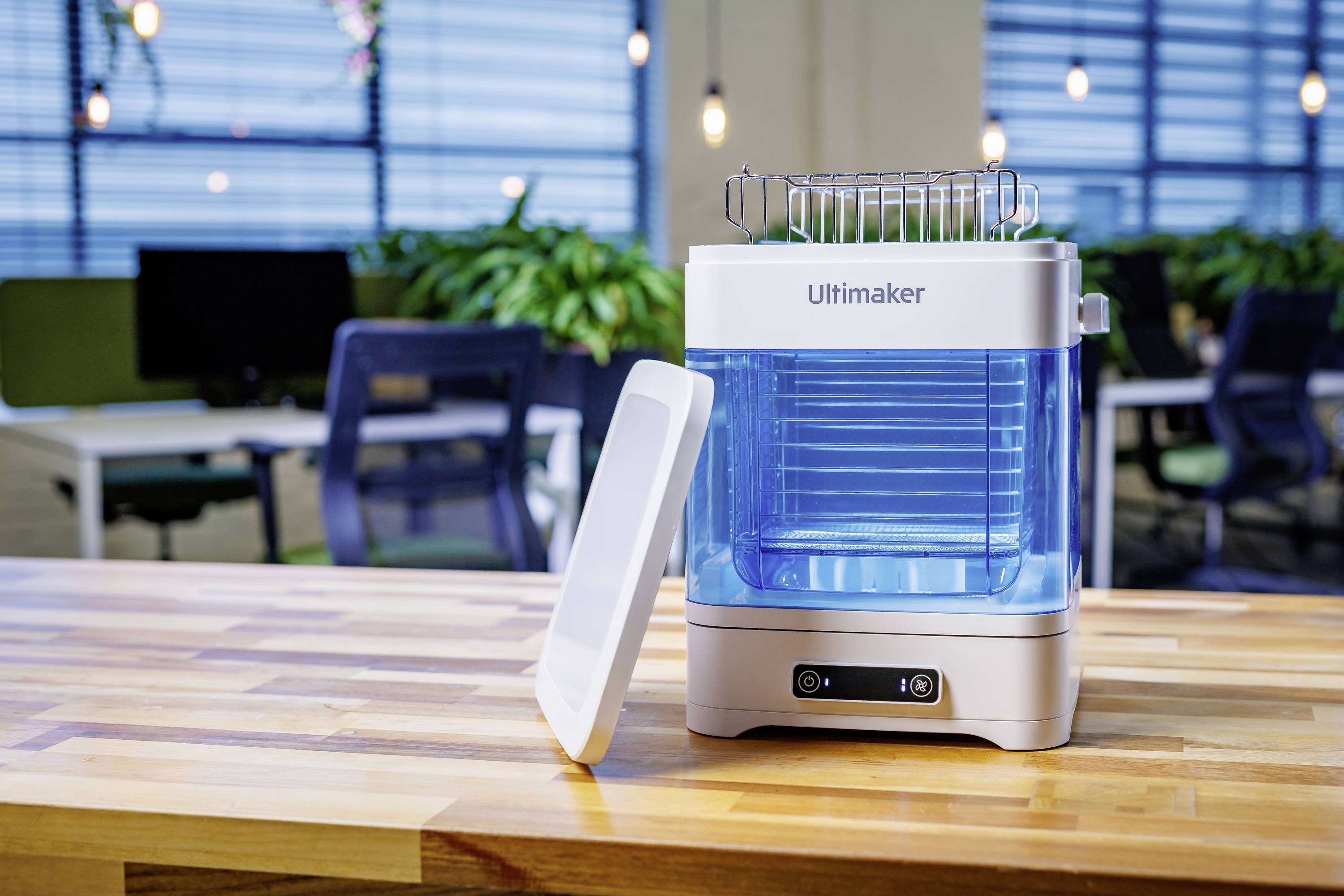 Een kleine, witte Ultimaker-luchtreiniger staat op een tafel in een modern kantoor met planten en computers op de achtergrond.