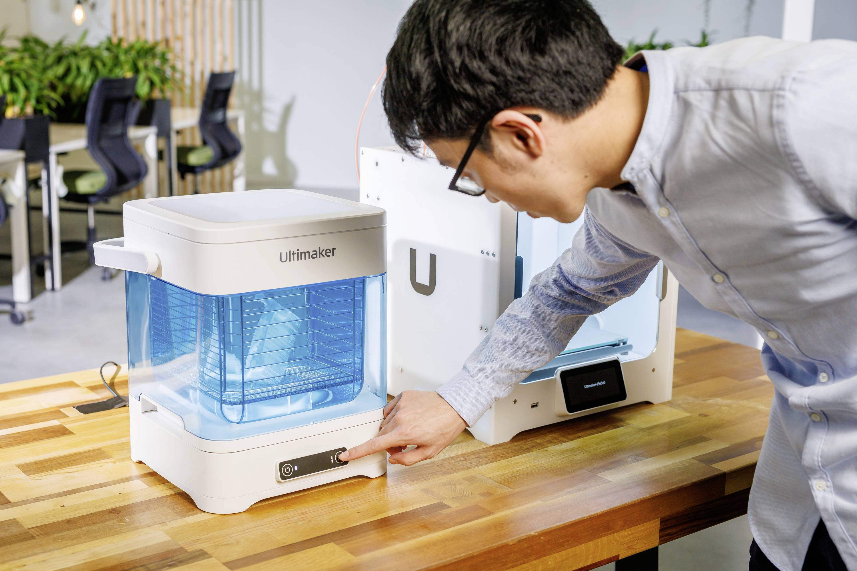 Een man bedient een 3D-printer op een houten tafel. De printer heeft een transparante blauwe kap. Op de achtergrond zijn kantoormeubels zichtbaar.