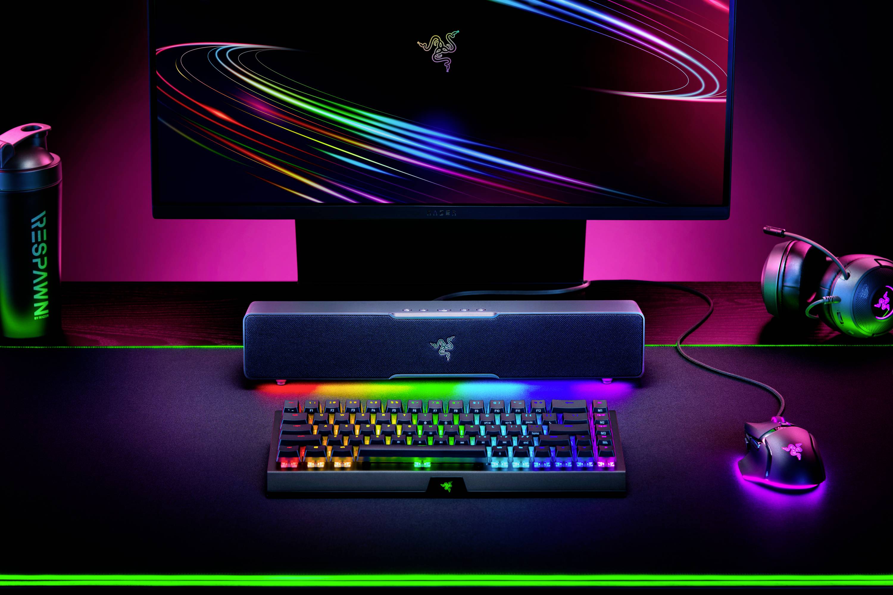 Een bureauopstelling met een verlicht toetsenbord, muis, speakers, monitor met Razer-logo en koptelefoon, allemaal gehuld in neonkleuren.