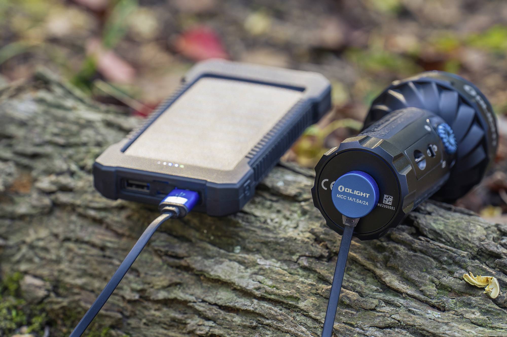 Powerbank en zaklamp op een houten boomstam in de buitenlucht, beide verbonden door een kabel.