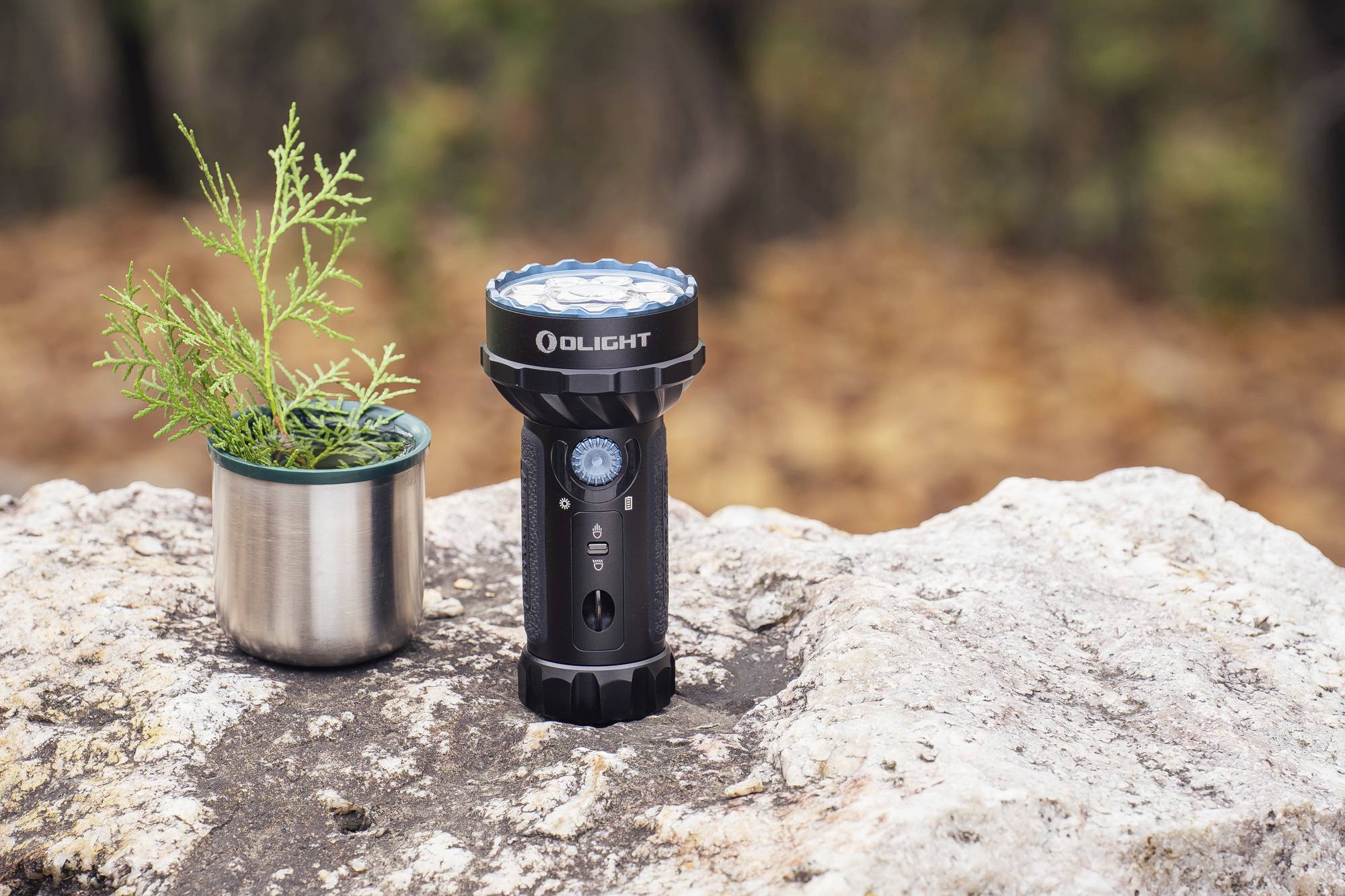 Een zwarte zaklamp staat op een steen naast een kleine, zilveren bloempot met groene takjes in de buitenlucht.