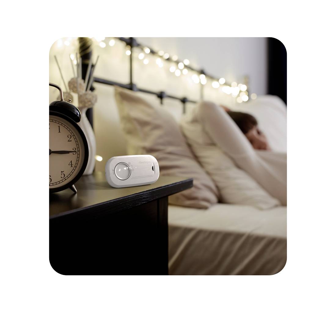 Een persoon rust ontspannen op een bed op de achtergrond, terwijl op de voorgrond een wekker en een elektronisch apparaat op een tafel staan.