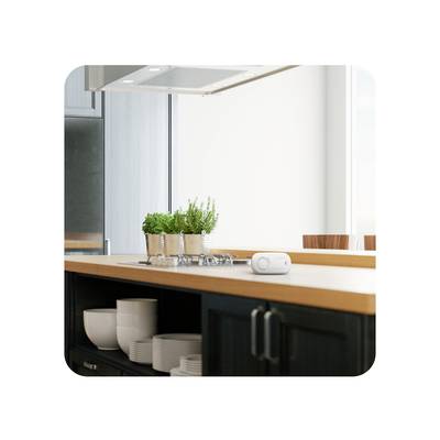 Een lichte keukenruimte met een fornuis op een houten werkblad. Kruidentopjes en een kleine witte luidspreker zijn zichtbaar.