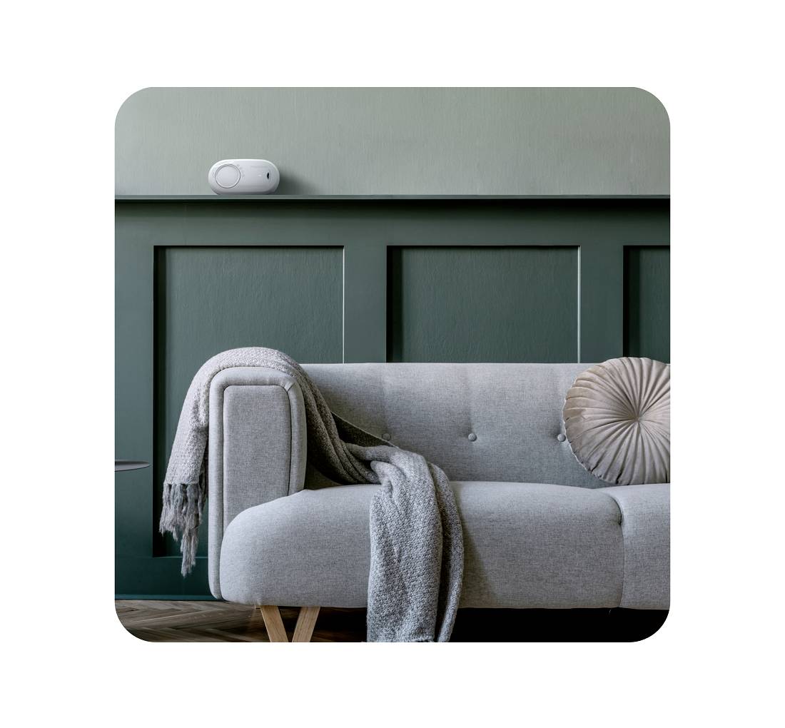 Een modern woonkamer met een grijze bank waarover een deken is gelegd, en een rond kussen. Aan de muur hangt een klein apparaat.