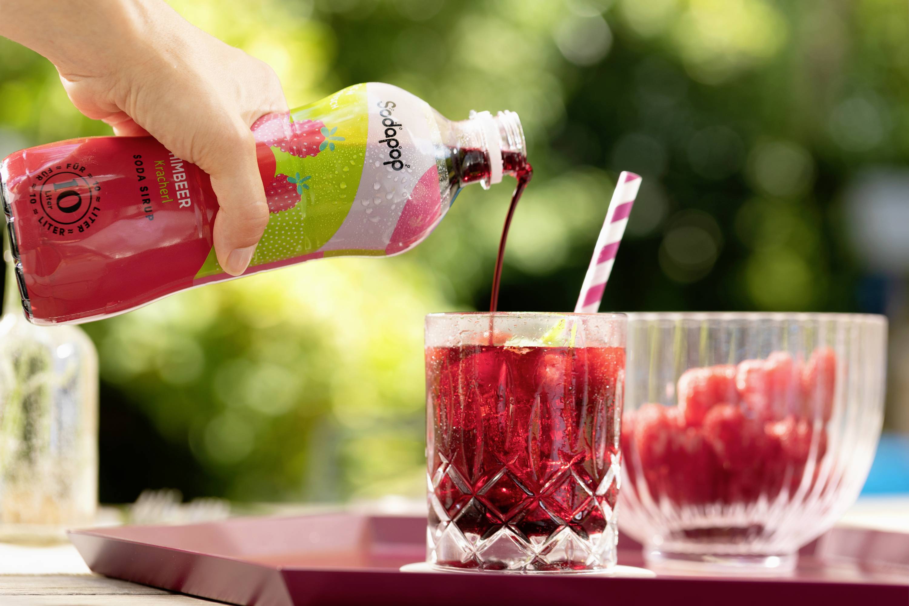 Een hand giet een rode drank uit een fles in een glas gevuld met ijsblokjes. Op de achtergrond zijn frambozen zichtbaar.
