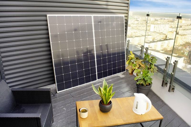Zonnepaneel op een balkon, naast planten. Een tafel met een waterkoker en koffiekopje toont huishoudelijk gebruik van hernieuwbare energie.