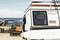 Een camper staat aan het strand met uitzicht op de zee. Een surfboard leunt tegen de zijkant, een oranje handdoek hangt naast de deur.
