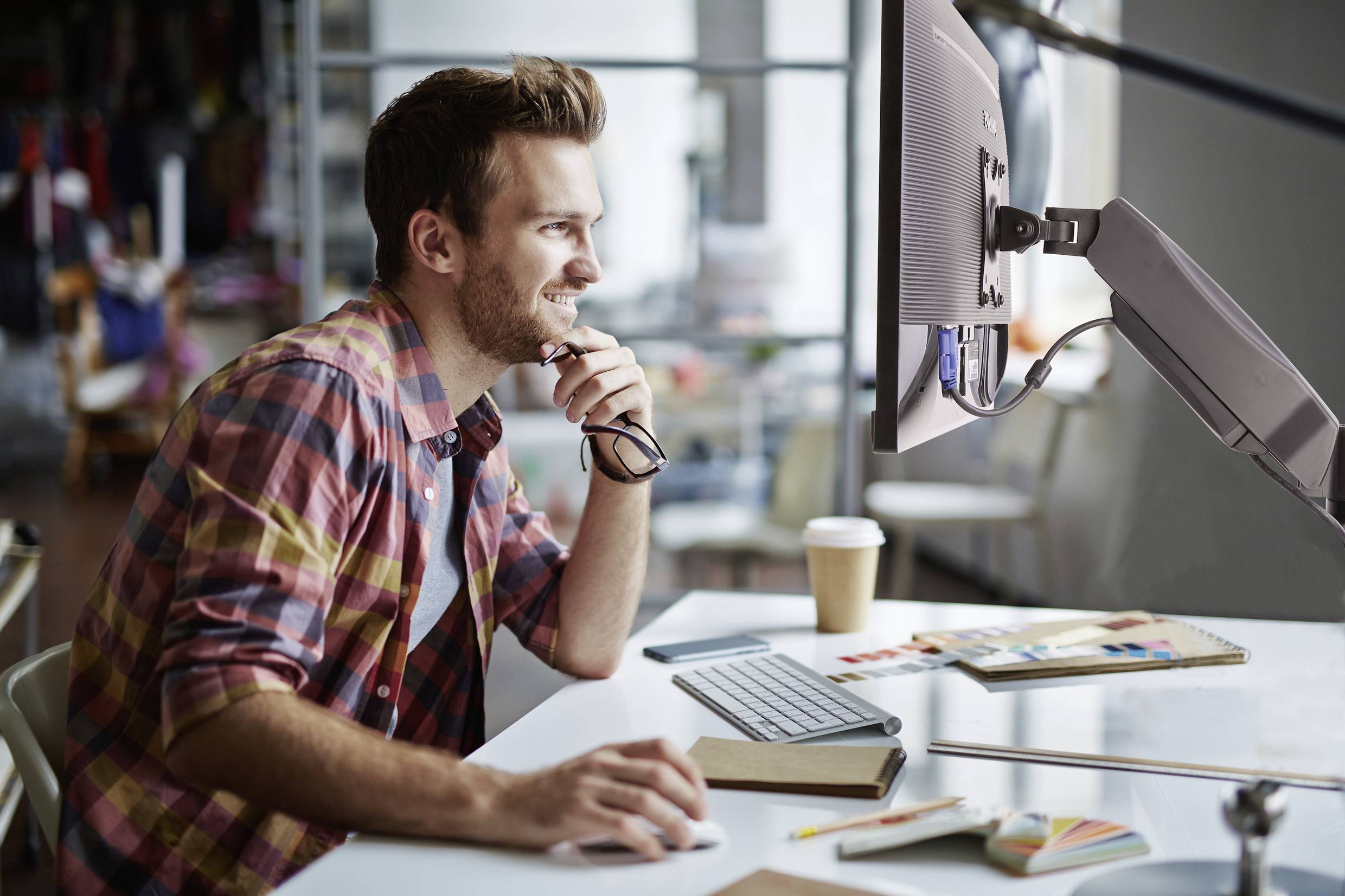 Een man zit glimlachend aan een bureau voor een computerscherm, omringd door notitieboeken en tekenmateriaal.
