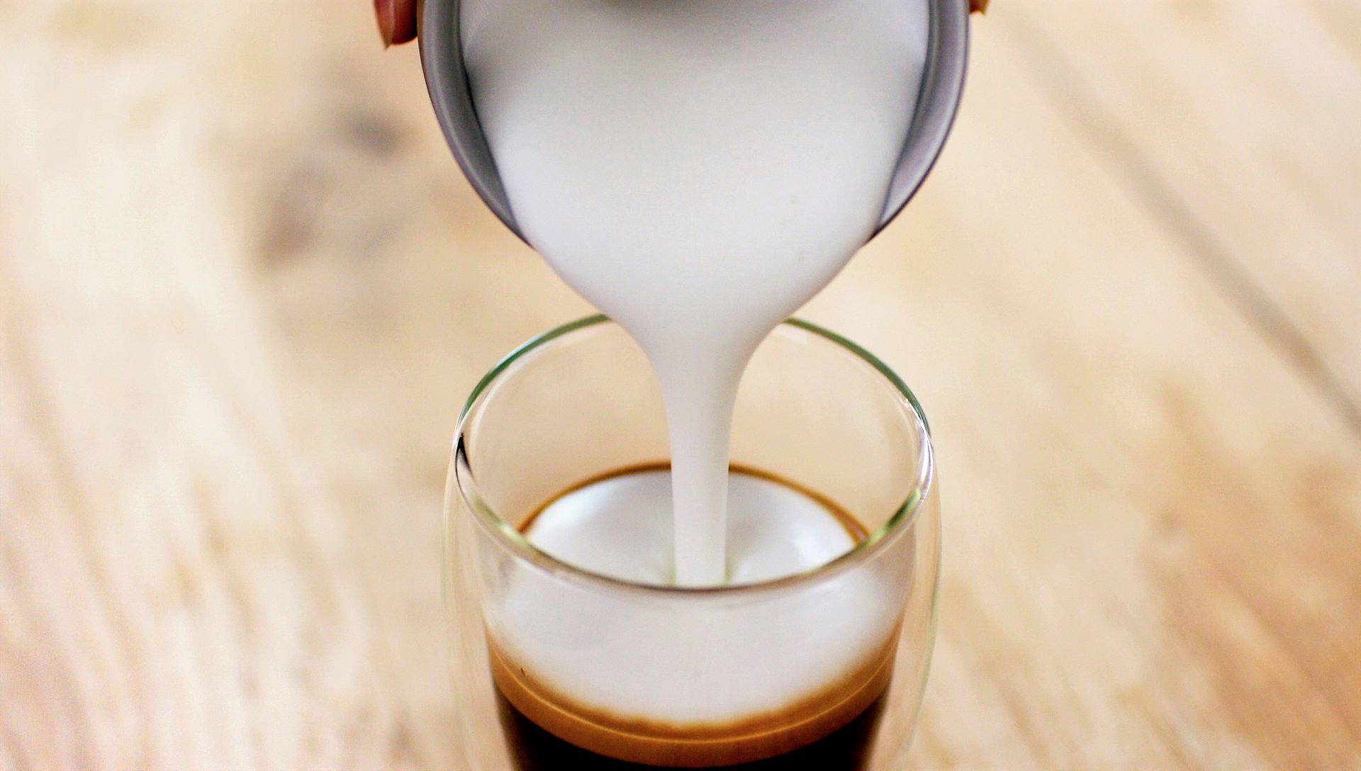 Een persoon giet opgeschuimde melk in een glas met espresso om een Latte Macchiato te bereiden, op een houten tafel.