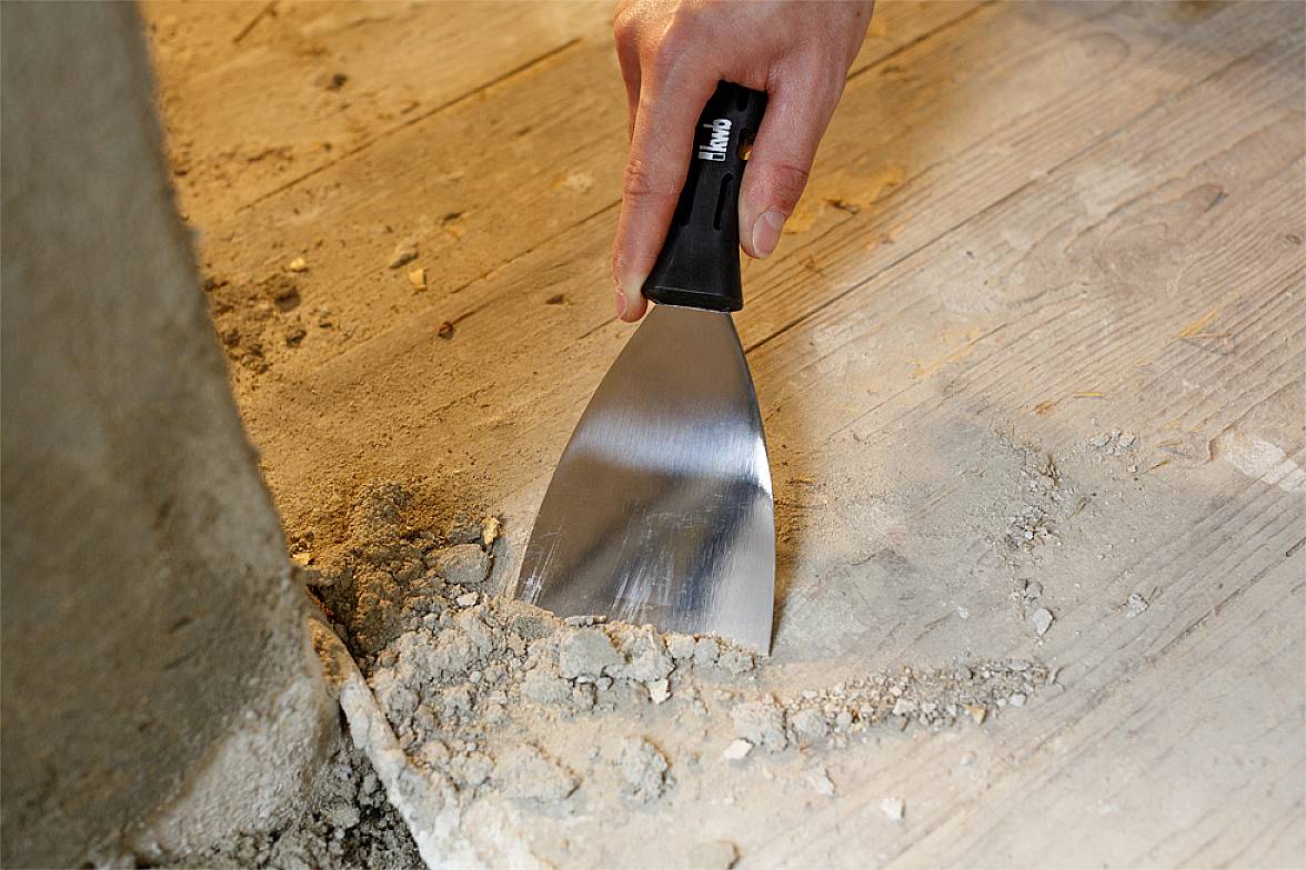 Een hand houdt een plamuurmes vast en verwijdert oude mortel van een houten vloer langs de rand van een betonnen plint.
