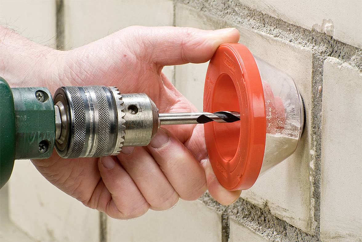 Een hand gebruikt een boormachine met een rode geleiding als sjabloon om een gat te boren in een witte bakstenen muur.