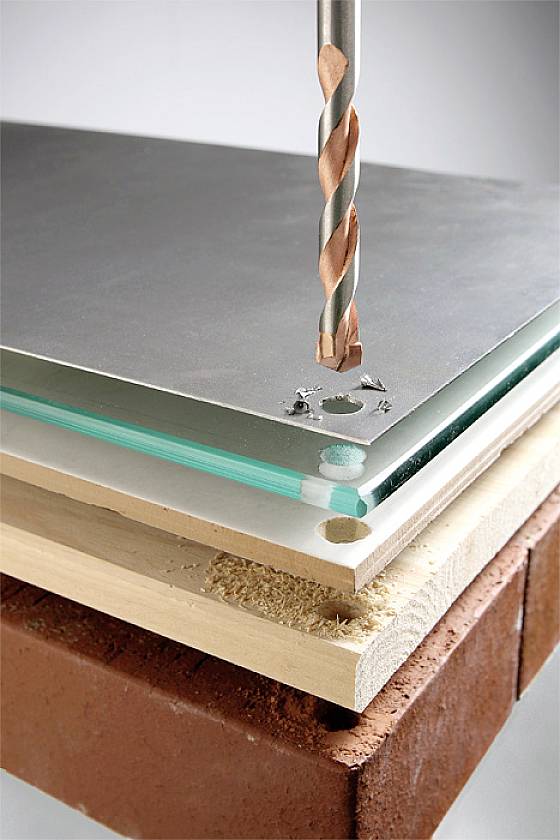 Een boormachine boort door een stapel uit verschillende materialen: metaalplaat, glas, hout en baksteen.