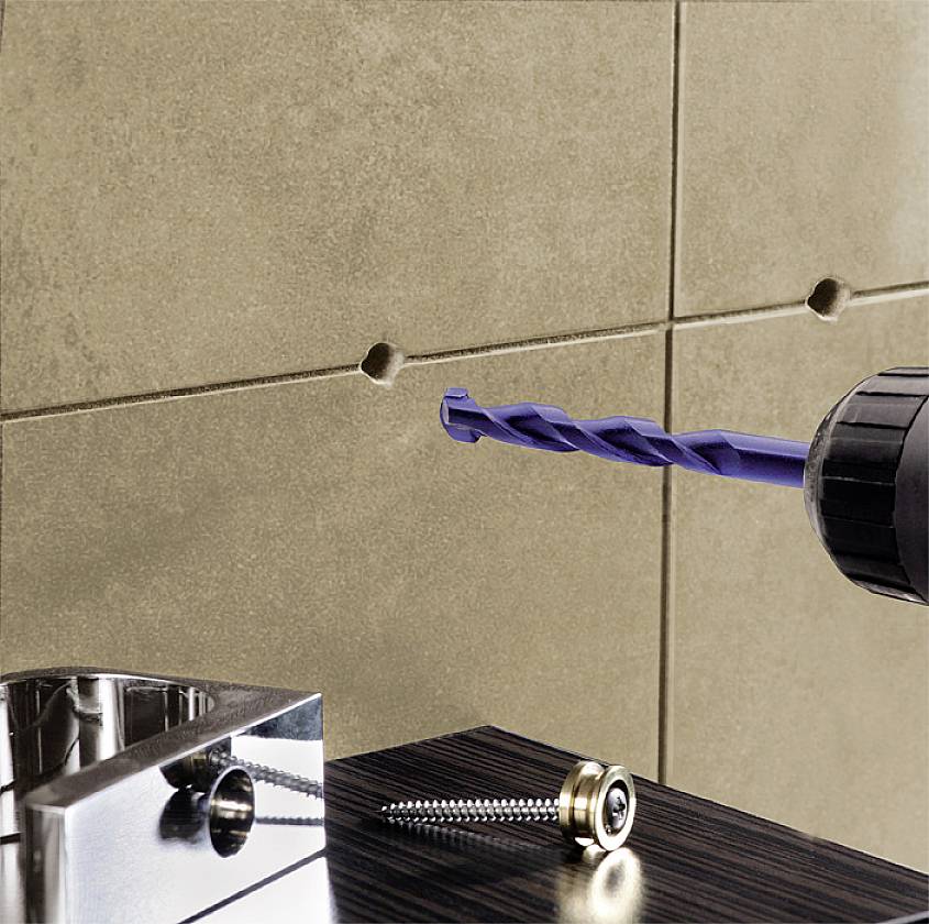 Een boormachine boort een gat in een lichte tegelmuur. Op een tafel op de voorgrond liggen een schroef en een metalen hoekprofiel.