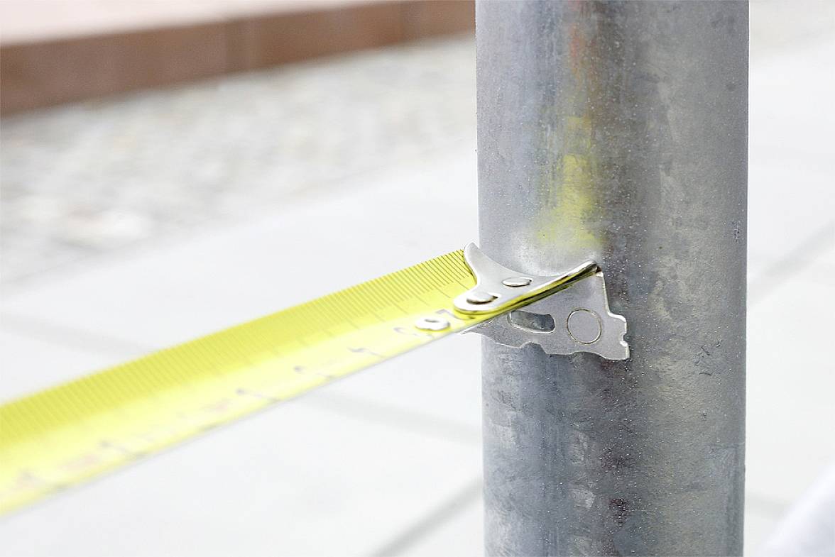 Een geel meetlint is gespannen rond een metalen paal om de omtrek te meten. Op de achtergrond is een onscherp voetpad zichtbaar.