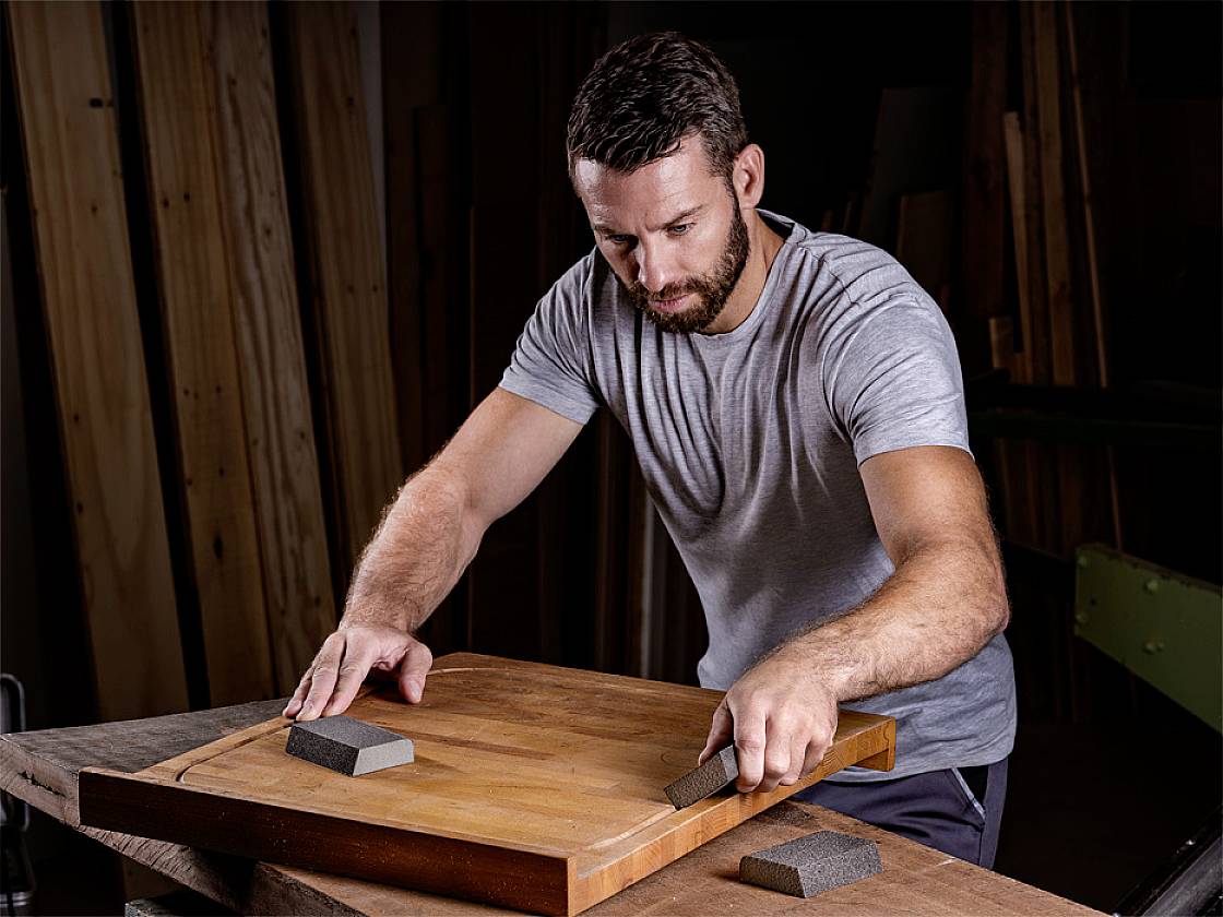 Een man schuurt een houten plank met schuurblokken in een werkplaats. Op de achtergrond zijn houten latten zichtbaar.