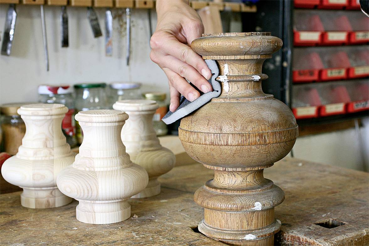 Een persoon schuurt een houten balk in een werkplaats. Op de achtergrond zijn verschillende onbewerkte stukken hout zichtbaar.