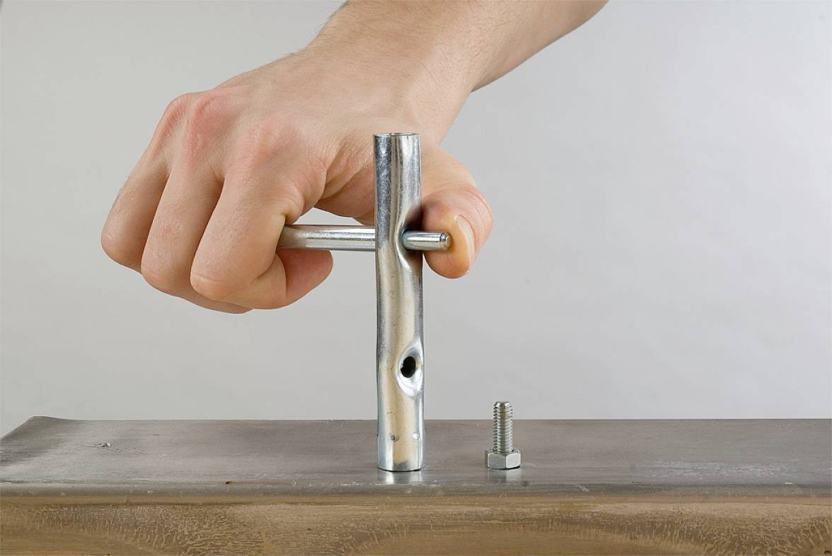 Een hand draait een schroef in een metalen buis op een tafel. Een moer ligt ernaast.
