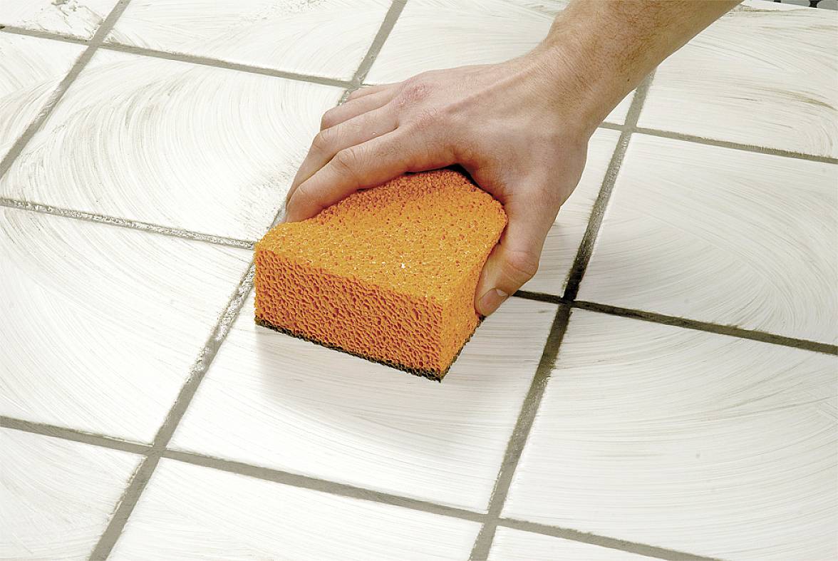 Een hand reinigt witte tegels met een oranje spons en verwijdert vuil uit de voegen.