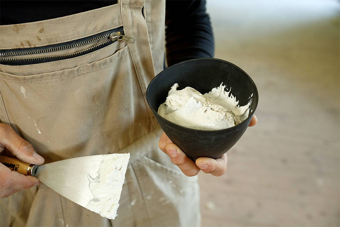 Een persoon houdt een pot en een pollepel met witte klei. De persoon draagt een bruine schort.