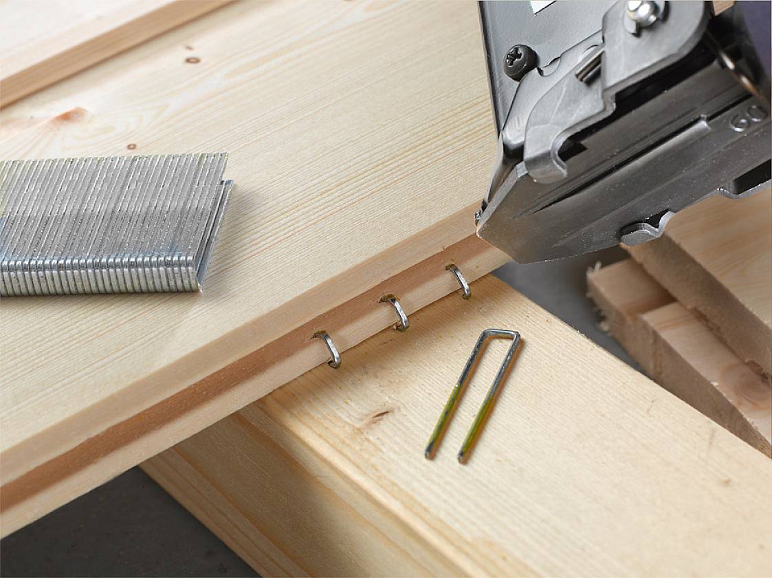 Een close-up van een nietmachine die metalen nietjes in hout aanbrengt. Rechts zijn losse nietjes zichtbaar.