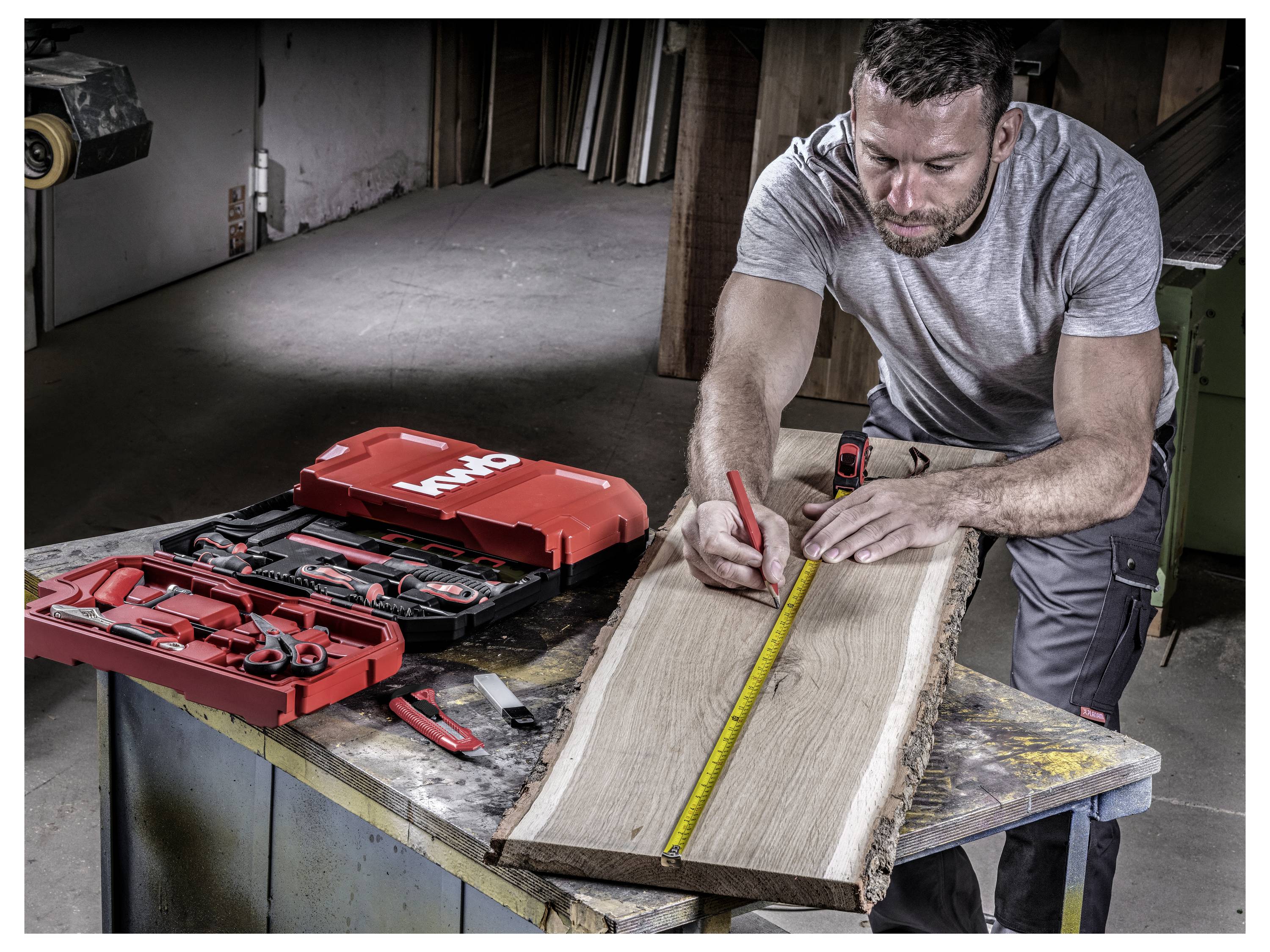 Een persoon meet een houten plank met een meetlint op een werkbanktafel. Een rode gereedschapskist met gereedschappen staat open naast hen.
