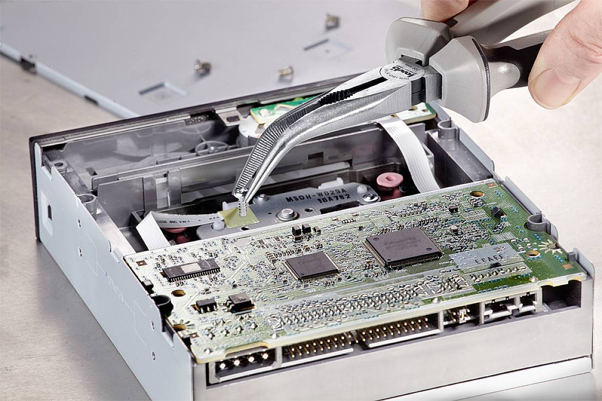 Een hand houdt een tang vast en werkt aan een opengehaald computerstation, waarbij de binnenstructuur en elektronische printplaat worden blootgesteld.
