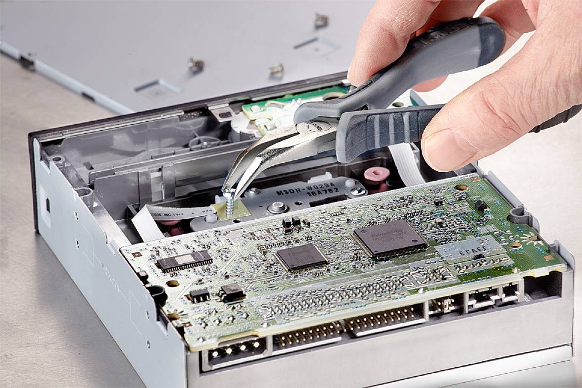 Een hand gebruikt een tang om te werken aan de open elektronica van een diskettestationnetje, wat laat zien hoe hardware wordt gerepareerd.