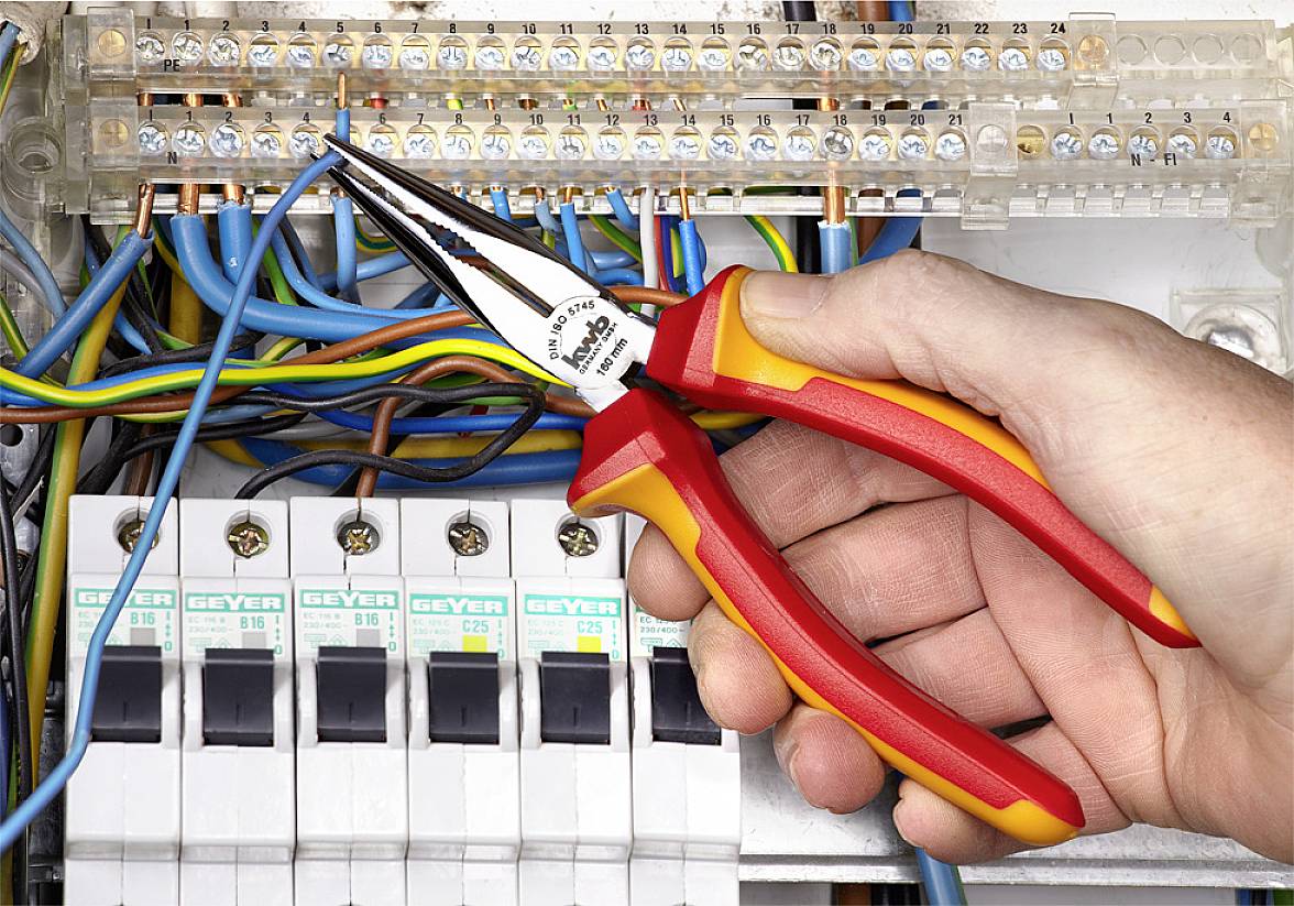 Een hand houdt een punttang vast en werkt aan kabels in een schakelkast, omgeven door gekleurde draden en zekeringautomaten.
