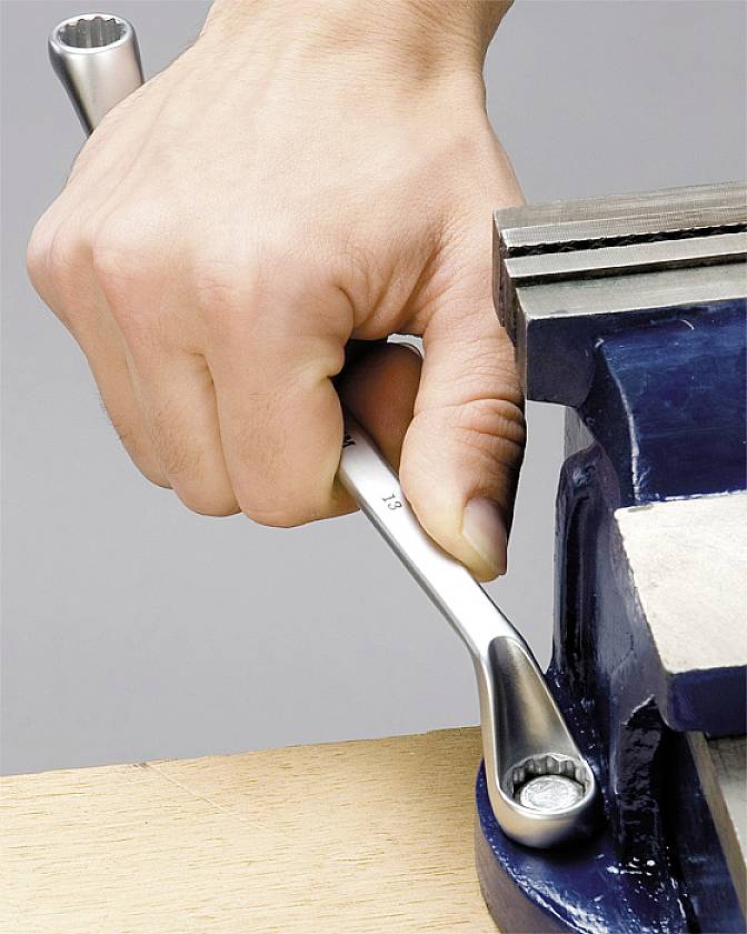 Een hand draait met een moersleutel een schroef aan die in een bankschroef is vastgeklemd.